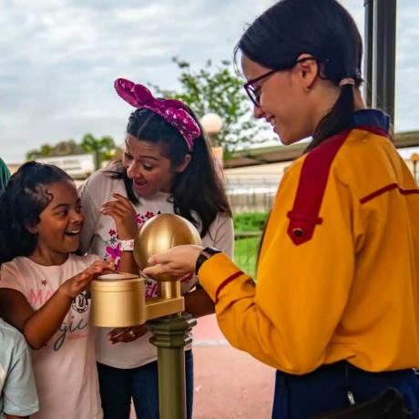 nasmijana djevojka lupka svojom čarobnom vrpcom i narukvicom po kiosku na ulazu u tematski park Walt Disney World, s majkom i članom glumačke ekipe koji joj uzvraćaju smiješak